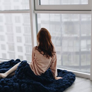 May include: A woman sits on a large, navy blue knitted blanket, looking out a window. She is wearing a light pink sweater and white socks.
