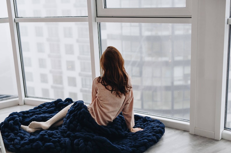 May include: A woman sits on a large, navy blue knitted blanket, looking out a window. She is wearing a light pink sweater and white socks.