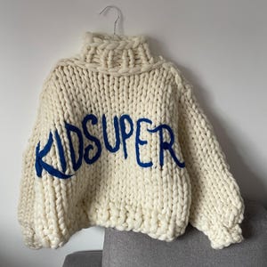 May include: Cream-colored, chunky knit sweater with a high turtleneck collar. The word "KIDSUPER" is embroidered across the front in blue. The sweater is hanging on a silver hanger against a white wall.