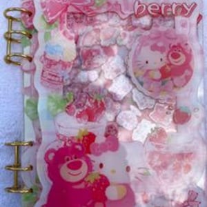 May include: A clear plastic binder with gold-tone metal rings featuring a pink and white theme. The cover displays the text "Strawberry" and illustrations of a pink bear and a white cat character, along with images of strawberries and bows.