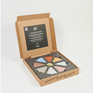 May include: A cardboard box with a lid open, revealing a circular display of eight small concrete samples in various colours. The box is labelled "CONCRETE SAMPLE SUPREME".