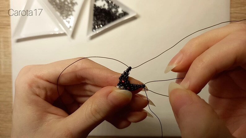 May include: A close-up of a person's hands working on a beaded jewelry project. The beads are black and red, and the thread is black. The person is holding the beads in their left hand and using their right hand to thread the beads.