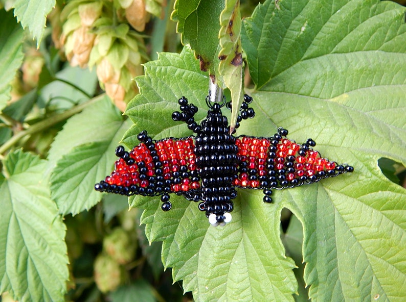 May include: A beaded bat charm with black and red beads. The bat is hanging from a green leaf.