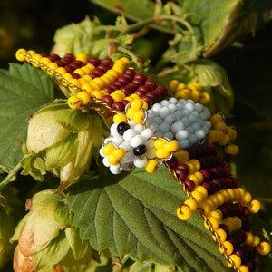 May include: A beaded bird figurine with white, yellow, and brown beads. The bird is perched on a green leaf with green and yellow flower buds.
