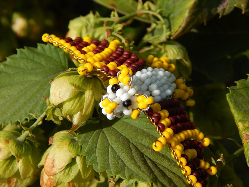 May include: A beaded bird figurine with white, yellow, and brown beads. The bird is perched on a green leaf with green and yellow flower buds.