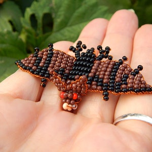 May include: A small beaded bat with brown and black beads, red eyes, and copper wire. The bat is held in a person's hand.
