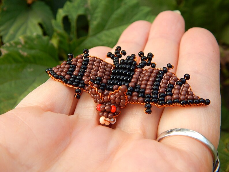 May include: A small beaded bat with brown and black beads, red eyes, and copper wire. The bat is held in a person's hand.