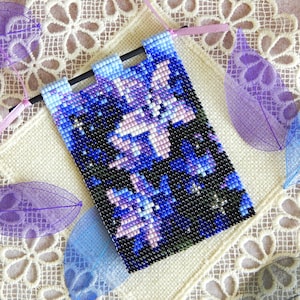 May include: A beaded wall hanging with a blue, purple, and white floral design. The hanging is attached to a wooden dowel rod with a pink ribbon.