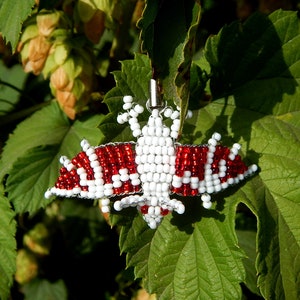 May include: A red and white beaded bat pendant hanging from a green leaf. The bat is made of small beads and has a wire frame.