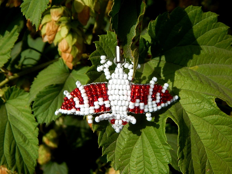 May include: A red and white beaded bat pendant hanging from a green leaf. The bat is made of small beads and has a wire frame.