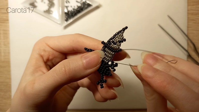 May include: A close-up of a beaded figure being made with black and silver beads. The figure is being held by a person's hands and is being worked on with a needle and thread.