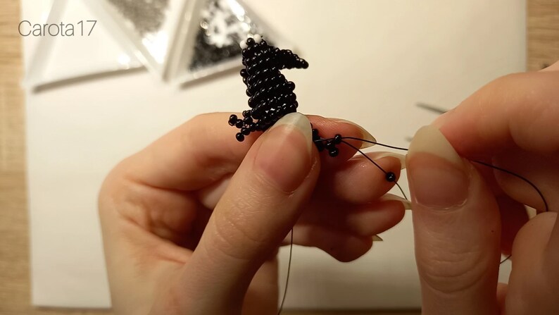 May include: A black beaded bird figurine is being strung with black thread. The bird has a long beak and a small body. The thread is being held by a hand with long, painted nails.