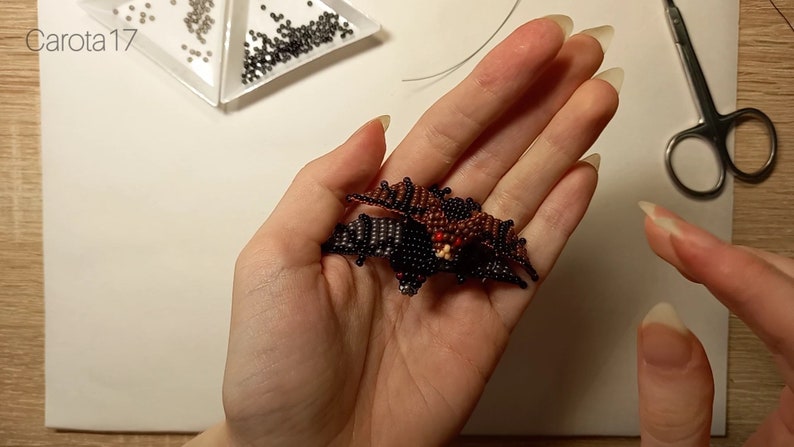 May include: A brown and black beaded bat with red eyes. The bat is held in a person's hand.