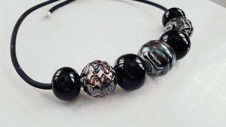 May include: A black elastic necklace with six beads. The beads are black, silver, and clear with a swirling pattern.