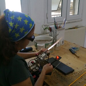 May include: A person wearing safety glasses and a blue bandana with yellow stars is using a torch to heat a glass rod. The person is working at a table with a variety of glass rods and tools.