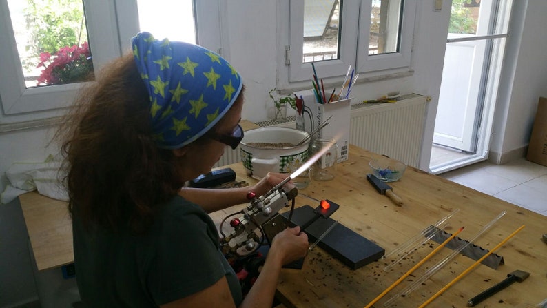 May include: A person wearing safety glasses and a blue bandana with yellow stars is using a torch to heat a glass rod. The person is working at a table with a variety of glass rods and tools.