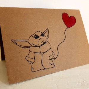 May include: A brown card with a black and white illustration of a character from the Star Wars franchise holding a red heart-shaped balloon.