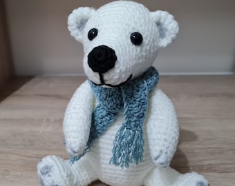 Può includere: Un orso polare all'uncinetto bianco con naso nero, occhi neri e una sciarpa con frange azzurra. L'orso presenta dettagli grigi sulle orecchie e sulle zampe. Il testo Little Green Bear è visibile nell'angolo in basso a sinistra.