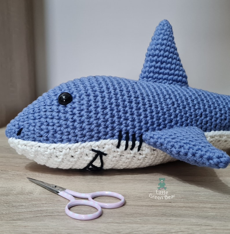 May include: A blue and white crocheted shark plush toy with a black plastic eye and stitched black gill details. Beside the shark lies a pair of small light pink scissors. The text Little Green Bear is printed in green near the scissors.