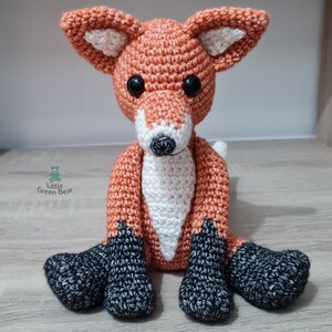 May include: A crocheted fox plush toy with orange and white fur and black feet. The fox is sitting with its legs crossed and its tail curled behind it.