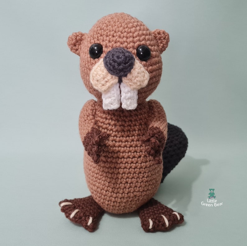 May include: A brown crocheted beaver with black eyes, a grey nose, and white teeth. The beaver is standing on a light blue background.
