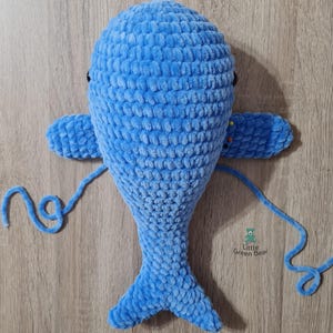 May include: A blue crocheted whale stuffed animal with black eyes and a tail. The whale is lying on a wooden surface with a blue yarn tail attached.