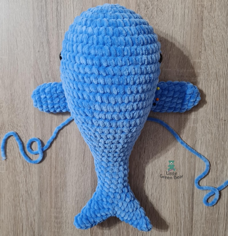 May include: A blue crocheted whale stuffed animal with black eyes and a tail. The whale is lying on a wooden surface with a blue yarn tail attached.