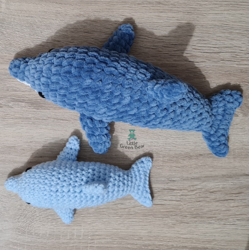 May include: Two crocheted dolphin plush toys made of soft yarn. The larger dolphin is a deep blue color, while the smaller dolphin is a light blue color. Both feature black bead eyes and a logo that reads Little Green Bear.