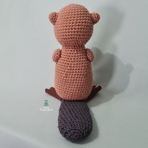 May include: A crocheted beaver toy with a brown body and a dark grey tail. The toy is sitting on a light green surface.
