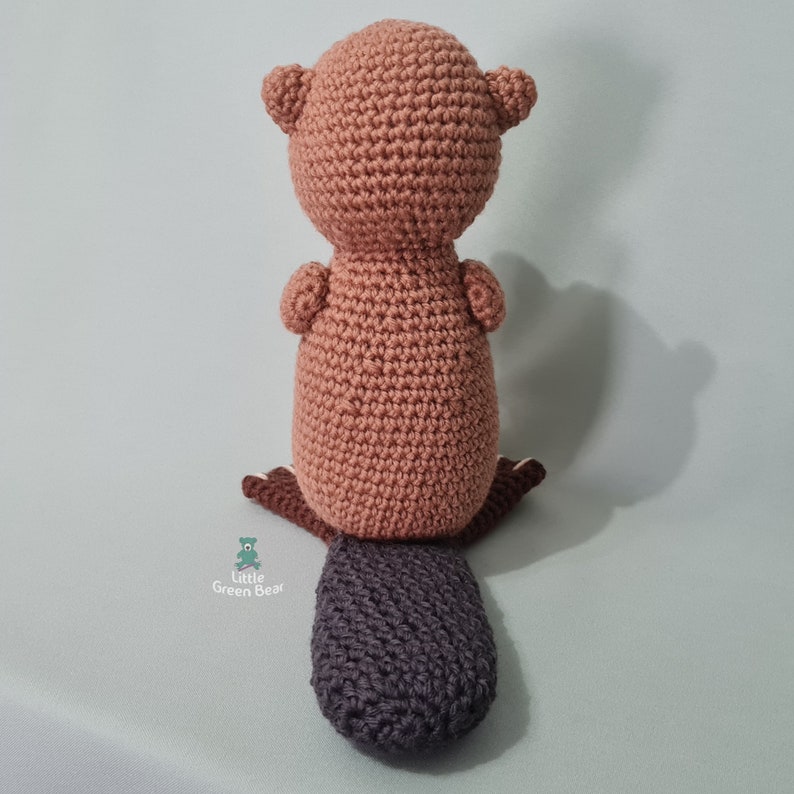 May include: A crocheted beaver toy with a brown body and a dark grey tail. The toy is sitting on a light green surface.