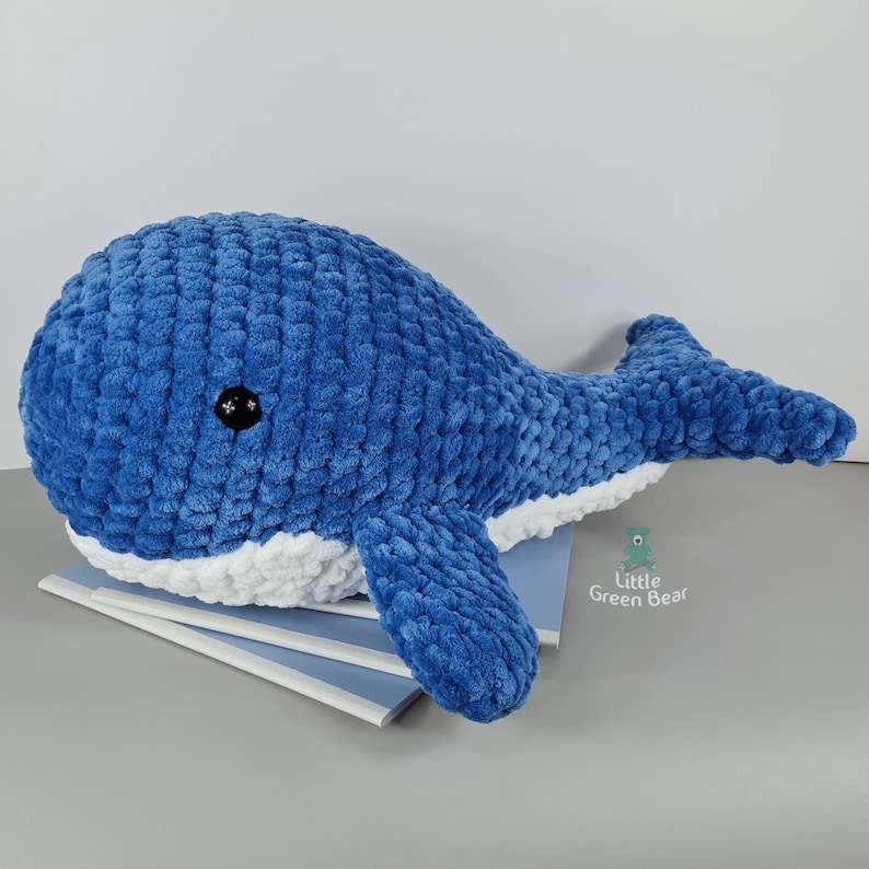 May include: A blue and white crocheted whale plush toy. The whale is lying on a stack of notebooks. The whale has black eyes and a white belly.