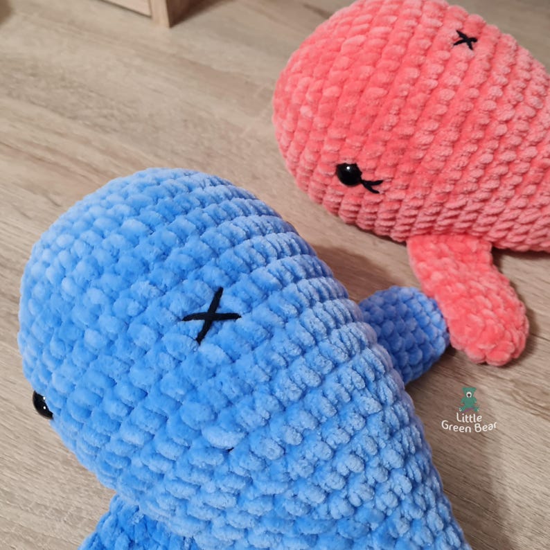 May include: Two crocheted whale plushies, one blue and one pink, with black embroidered eyes and a black X on the back. The plushies are made with a textured yarn and have a soft, cuddly appearance.