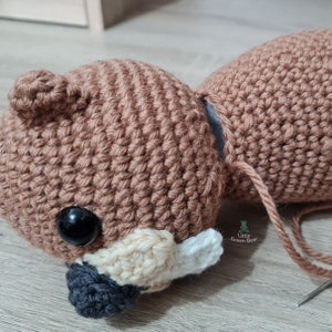 May include: A brown crocheted bear head with black eyes, a white nose, and a black mouth. The bear head is being sewn together with brown yarn.
