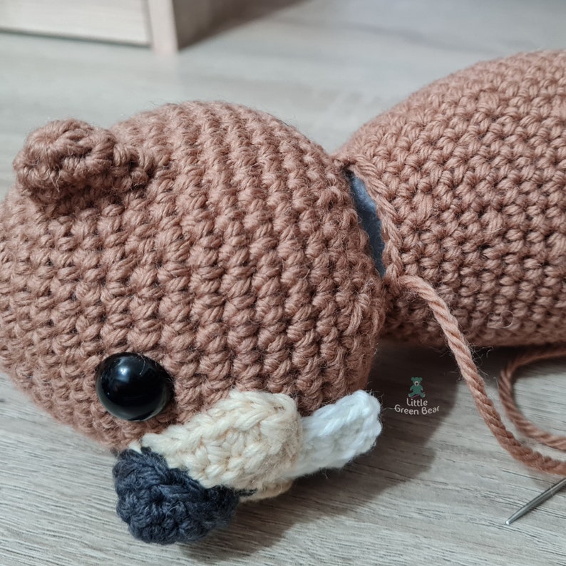 May include: A brown crocheted bear head with black eyes, a white nose, and a black mouth. The bear head is being sewn together with brown yarn.