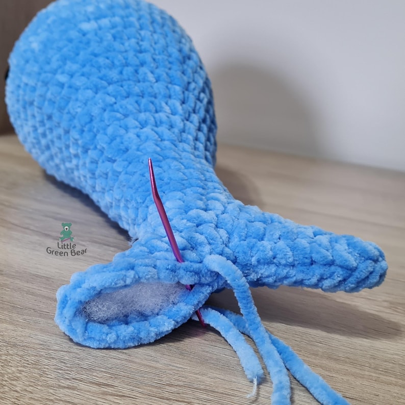 May include: A blue crocheted whale tail with a pink crochet hook and white stuffing inside. The tail is being sewn closed with a blue yarn.