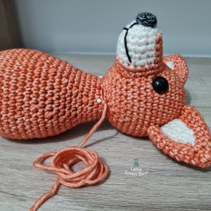 May include: A crocheted fox toy with orange and white fur, being sewn together with orange thread. The fox has black eyes and a black nose.