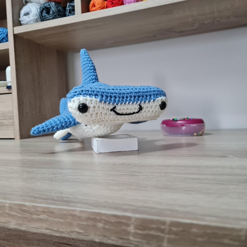 May include: A crocheted hammerhead shark plush toy featuring a blue top and white underside. The toy has black plastic safety eyes and a stitched black mouth. It is balanced on a small white rectangular block, showcasing its distinct head shape.