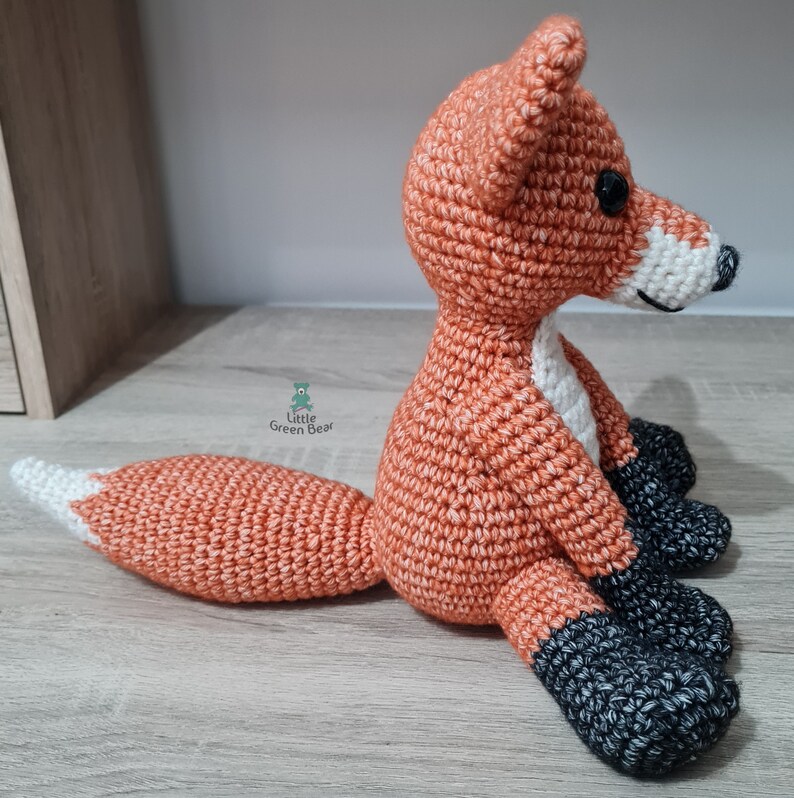 May include: A crocheted fox plush toy with orange and white fur, black paws, and a white tip on its tail. The fox is sitting on a wooden surface.