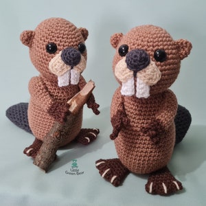 May include: Two brown crocheted beaver plushies, one holding a small branch. The beavers have black eyes, white teeth, and brown noses. The plushies are sitting on a light green surface. The text "Little Green Bear" is visible in the bottom centre of the image.