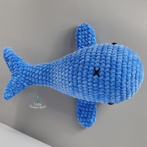 May include: A blue crocheted whale plush toy with black embroidered eyes and a stitched mouth. The whale is lying on its side with its tail in the air.