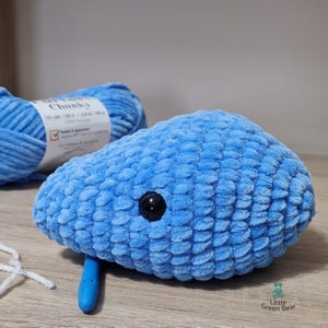 May include: A blue crocheted whale plushie with a black button eye. The whale is made with chunky yarn and has a textured surface.