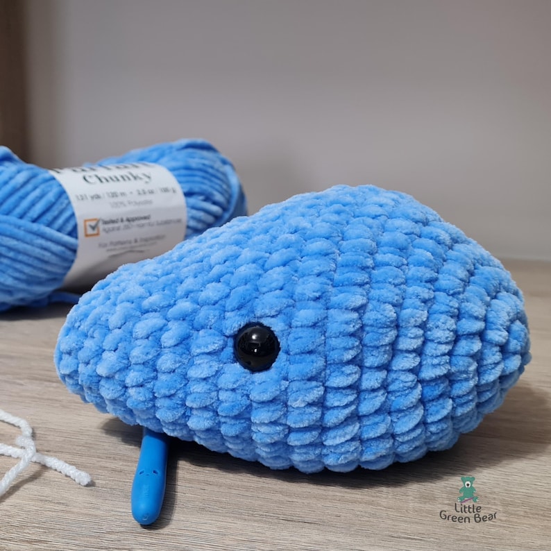 May include: A blue crocheted whale plushie with a black button eye. The whale is made with chunky yarn and has a textured surface.