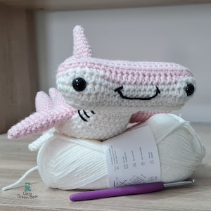 May include: A crocheted hammerhead shark toy in light pink and white yarn with black safety eyes and a stitched mouth. It rests on a ball of white yarn with a care label and a purple crochet hook. The logo Little Green Bear is visible.
