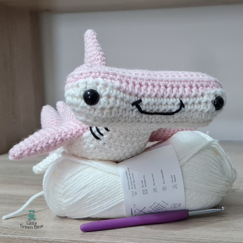 May include: A crocheted hammerhead shark toy in light pink and white yarn with black safety eyes and a stitched mouth. It rests on a ball of white yarn with a care label and a purple crochet hook. The logo Little Green Bear is visible.