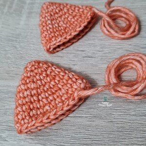 May include: Two crocheted triangle shapes in a peach color with white yarn accents. The shapes have long tails of yarn attached to them.