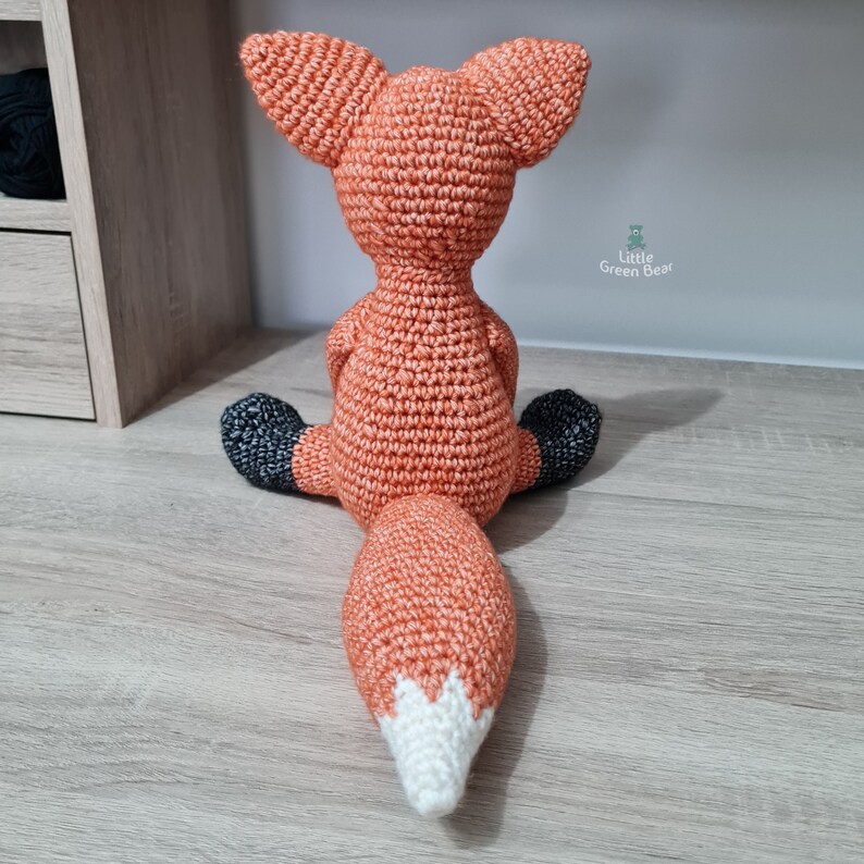 May include: A crocheted fox plush toy with a white tipped tail. The fox is sitting on a wooden surface.