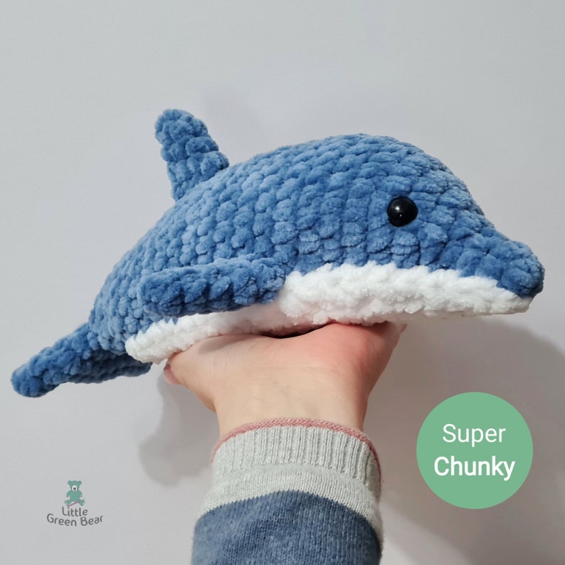 May include: A blue and white crocheted dolphin plush toy with a textured, chunky yarn finish and black plastic eyes. The toy features a dorsal fin, side fins, and a tail. A green circular graphic on the image contains the text Super Chunky.