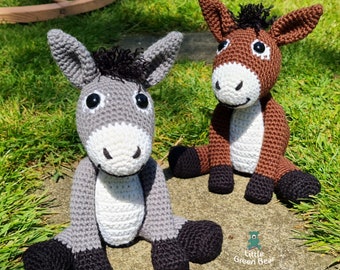May include: Two crocheted donkey toys, one grey and one brown, sitting on a stone path with green grass in the background. The grey donkey has black hooves and a black mane. The brown donkey has black hooves and a black mane. The text "Little Green Bear" is visible in the bottom right corner of the image.