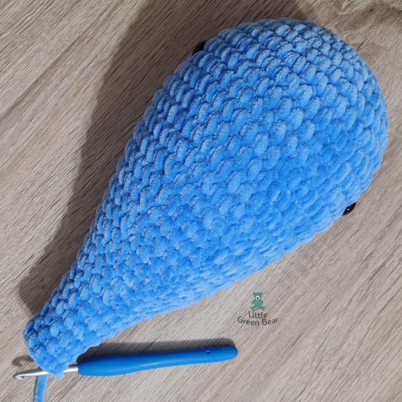 May include: A light blue crocheted whale tail with black eyes. The tail is lying on a wooden surface with a blue crochet hook next to it.
