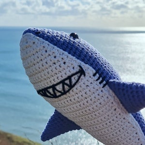 May include: A blue and white crocheted shark plush toy featuring a stitched black mouth with visible teeth and a small black bead eye. The shark is crafted with a textured yarn pattern, showing a white underbelly and a blue upper body.